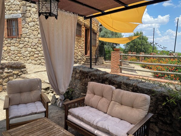 Un área de estar al aire libre con dos sillones acolchados y un sofá, una pared de piedra, una columna de ladrillo y un dosel amarillo.