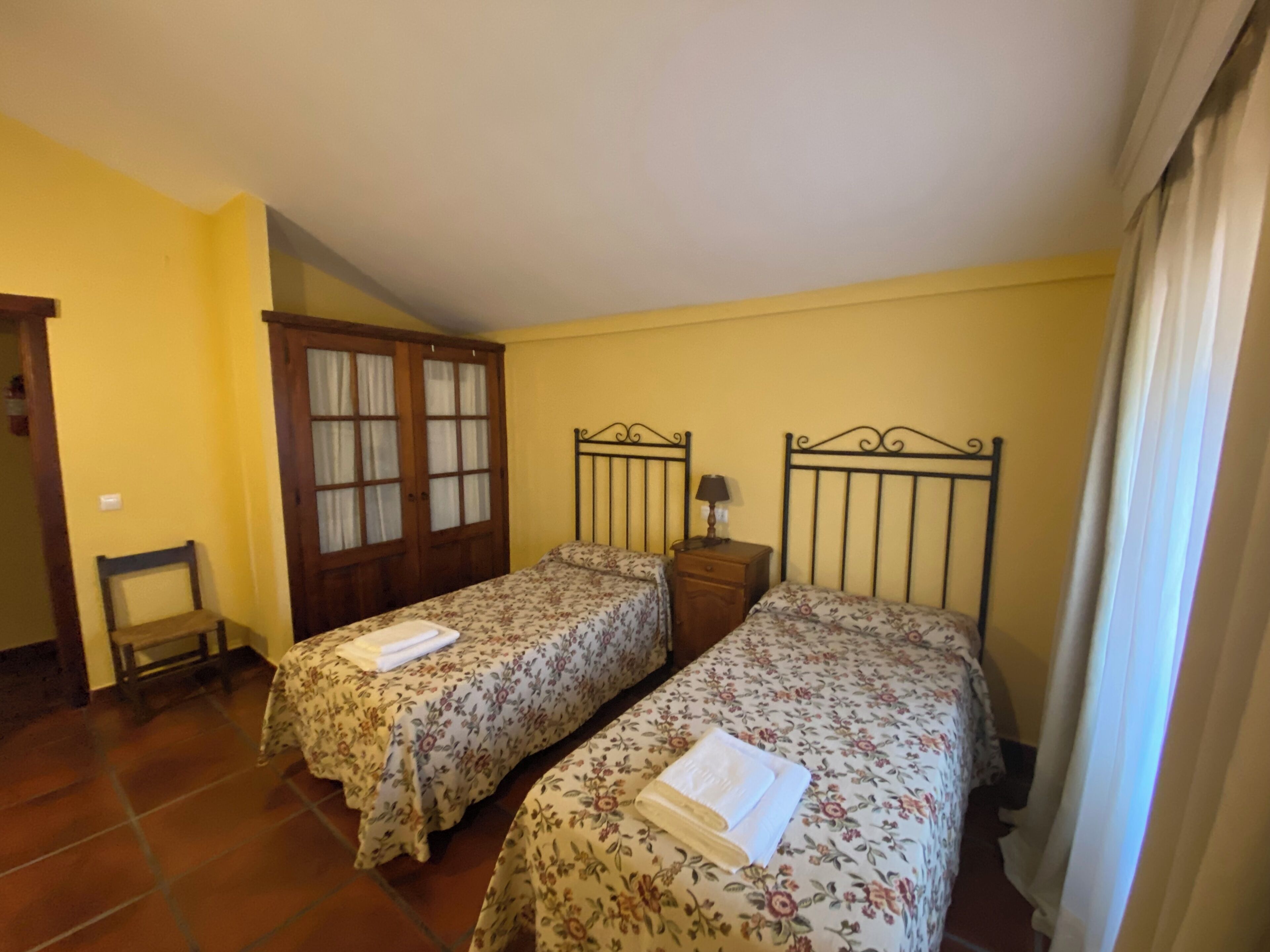 Una habitación con dos camas, un armario de madera, una silla pequeña y una mesita de noche con una lámpara.