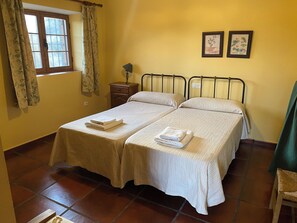 Cama doble con dos almohadas y toallas dobladas, una mesita de noche con lámpara, una ventana con cortinas y dos cuadros enmarcados en la pared.