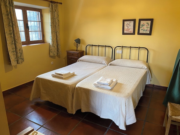 A double bed with two pillows and folded towels, a bedside table with a lamp, a window with curtains, and two framed pictures on the wall.