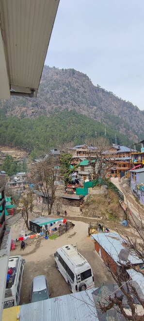 A mountainous landscape with a village. There are several buildings, some with green roofs, and a few with blue roofs. A white bus is parked in the foreground. - Woodward 2 by 4wildflower (Bhuntar)
