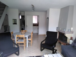 Un espacio habitable compacto con una mesa de comedor, sillas, un sillón negro, una chimenea y una zona de cocina al fondo.