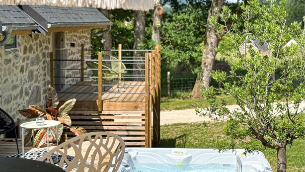 Un jacuzzi con una terraza de madera, un edificio de piedra con techo de paja y un pequeño árbol.