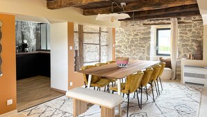 Comedor con una mesa de madera, sillas amarillas y un banco. Se observa una pared de piedra y una ventana con cortinas.