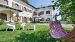 A courtyard with outdoor seating, a hammock, and a building with arched windows and balconies. - Room 'Agriturismo Cascina Favorita - D3' with Shared Garden and Wi-Fi (Castelletto Merli)