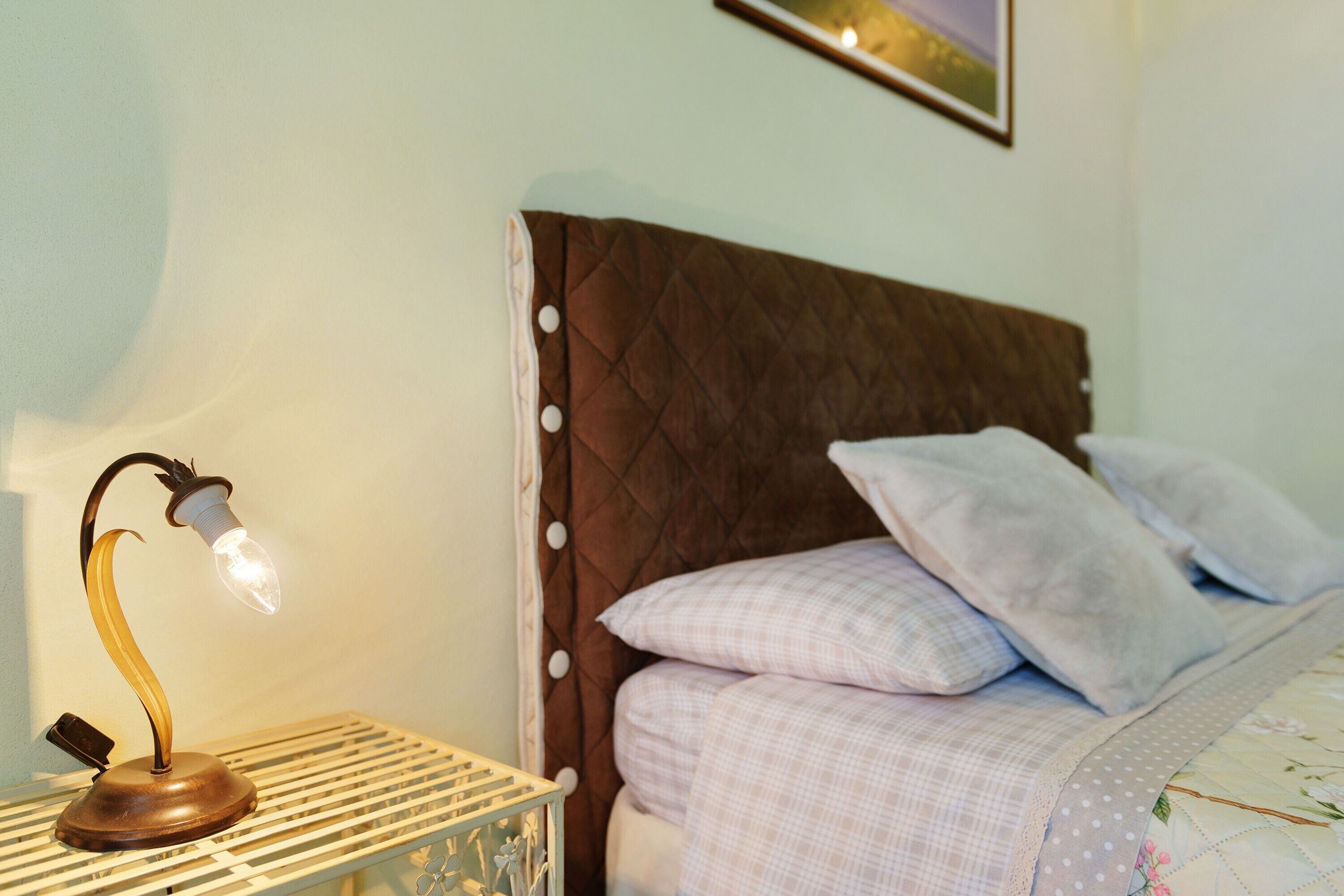 Een bed met een geborduurde hoofdbord, een nachtkastje met een lamp, en een aan de muur hangend schilderij.