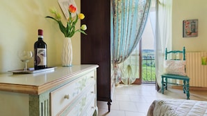 A bedroom with a bed, a dresser, a vase with flowers, a wine bottle, and a view of the outdoors. - Room 'Agriturismo Cascina Favorita - D3' with Shared Garden and Wi-Fi (Castelletto Merli)