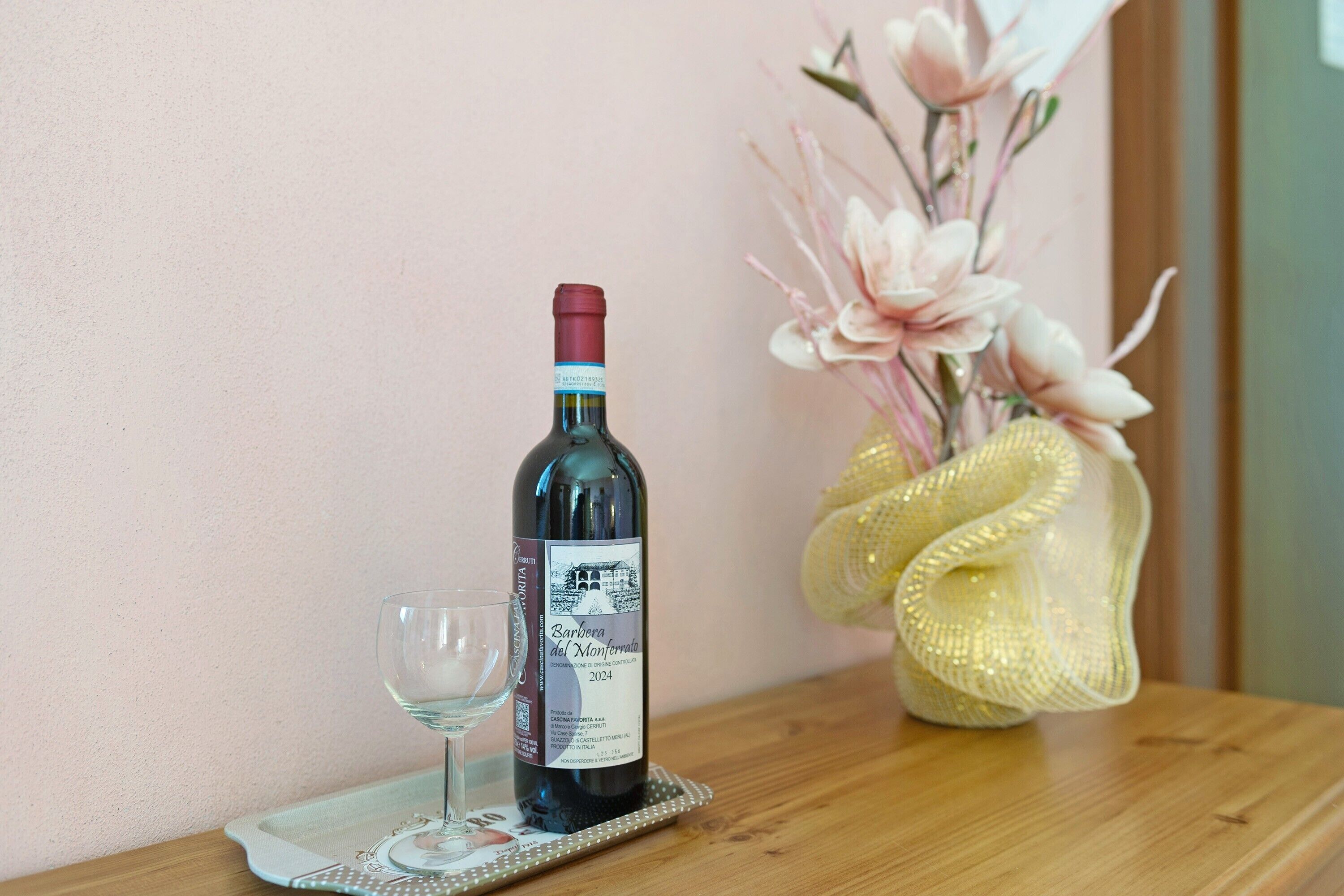 Eine Weinflasche auf einem Tablett mit einem Glas, eine dekorative Vase mit Blumen und ein Holztisch an einer rosafarbenen Wand.