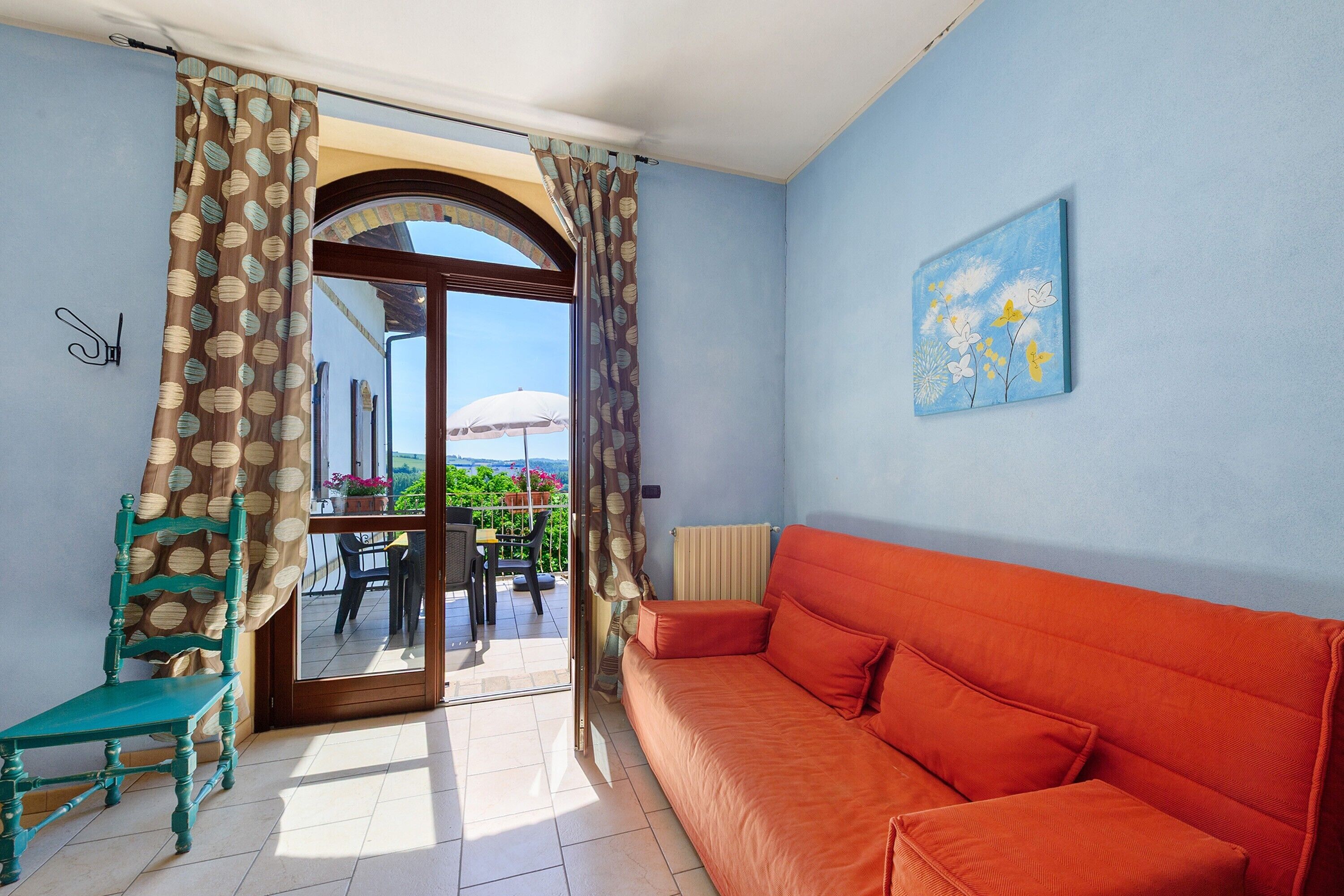 A room with a sofa, a chair, a painting, and a view through a glass door to an outdoor area with a table and chairs.