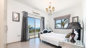 A bedroom with a bed, a nightstand, a lamp, a window with a view, and a chandelier. - Ayia Napa Panorama Villa (Ayia Napa)