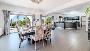 A spacious dining area with a large table and chairs, a kitchen with a central island, and a view of greenery through the glass doors. - Ayia Napa Panorama Villa (Ayia Napa)