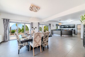 A spacious dining area with a large table and chairs, a kitchen with a central island, and a view of greenery through the glass doors.