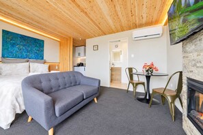 A modern hotel room with a grey sofa, a dining table with chairs, a fireplace, and a large TV mounted on the wall.