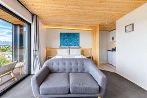 A modern bedroom with a large bed, a grey upholstered armchair, a wooden ceiling, and a view of the outdoors.