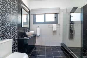 Une salle de bain moderne avec un grand miroir, un lavabo blanc, un plan de toilette noir, une douche en verre et une fenêtre au-dessus du lavabo.