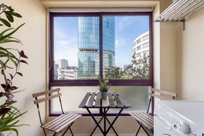 Un balcon donnant sur des immeubles modernes, une table en bois avec des chaises et une plante en pot.