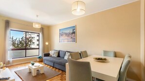 A modern living room with a sofa, dining table, and chairs, large windows with a view, and a hanging light fixture. - Apartment 'Destiny Oporto' with Wi-Fi (Maia)