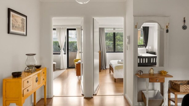 Una habitación de hotel moderna con suelo de madera, una cama con ropa de cama blanca, una mesita con una cesta y un espejo.