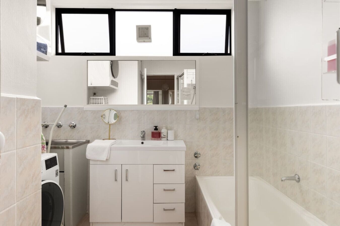 Ein modernes Badezimmer mit integrierter Waschmaschine, einem Waschtisch mit Schubladen, einem Spiegel und einer Badewanne.