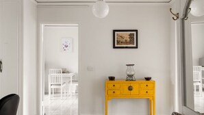 Una habitación con una puerta blanca, un aparador amarillo con cajones, un cuadro enmarcado en la pared y una silla blanca al fondo.