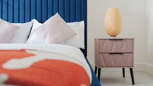 Een net opgemaakt bed met een blauw hoofdbord, een bedlampje en een nachtkastje met lades.
