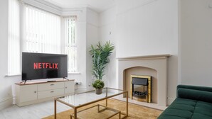 Een woonkamer met een televisie op een houten kast, een groene bank, een glazen salontafel, een potplant en een open haard.