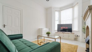Een woonkamer met een groene bank, een glazen salontafel, een televisie waarop 'NETFLIX' wordt weergegeven en een raam met jaloezieën.