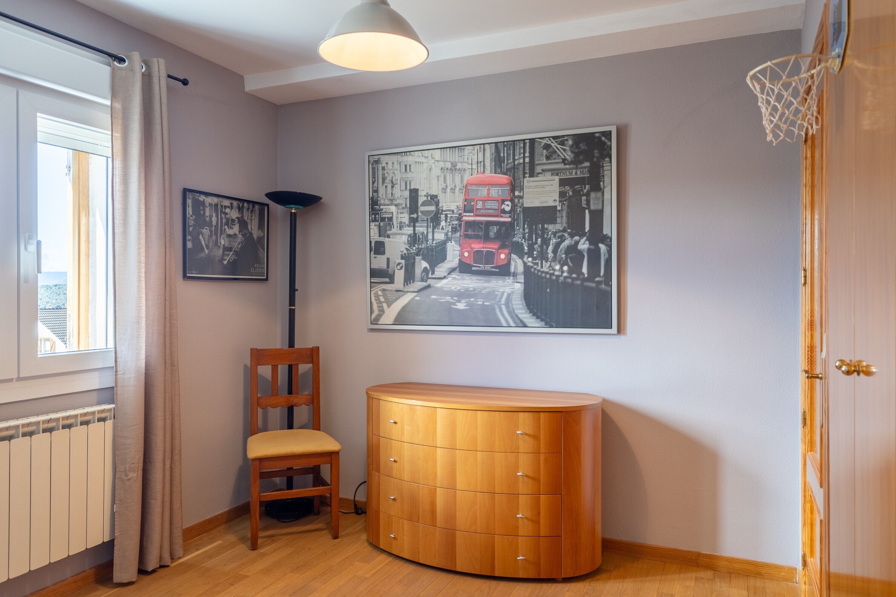 Una habitación con un armario de madera, una silla, un cuadro con un autobús de dos pisos y un aro de baloncesto.