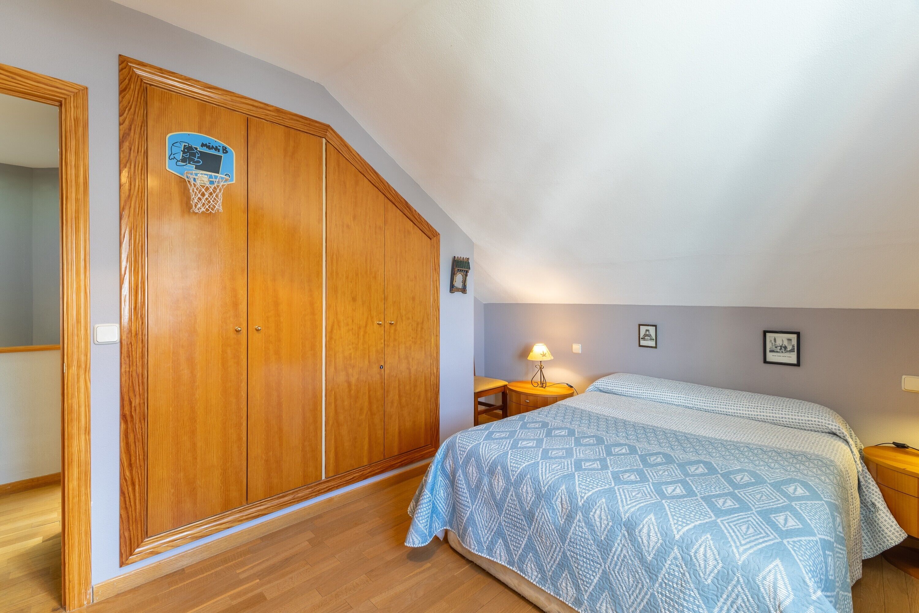 Un dormitorio con un amplio armario de madera, una cama con colcha estampada, una mesita de noche con lámpara y un aro de baloncesto instalado en la pared.