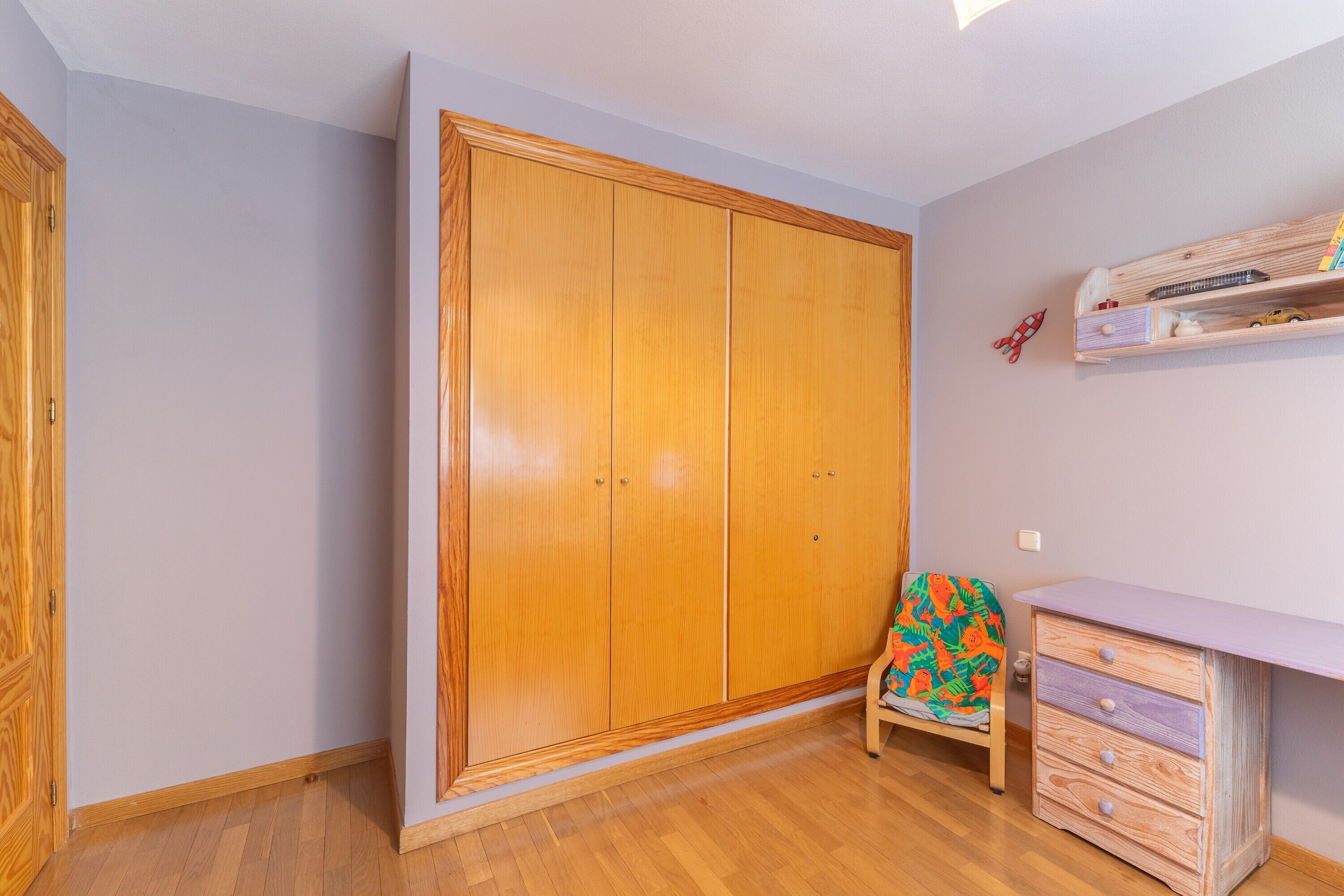 Una habitación con un armario de madera, una pequeña silla de madera con funda colorida y un aparador de madera con cajones.