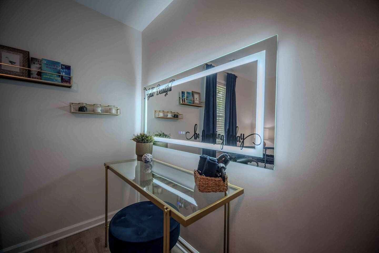 Una stanza con un tavolo in vetro, un pouf blu, uno specchio con cornice decorativa e mensole con libri e oggetti decorativi.