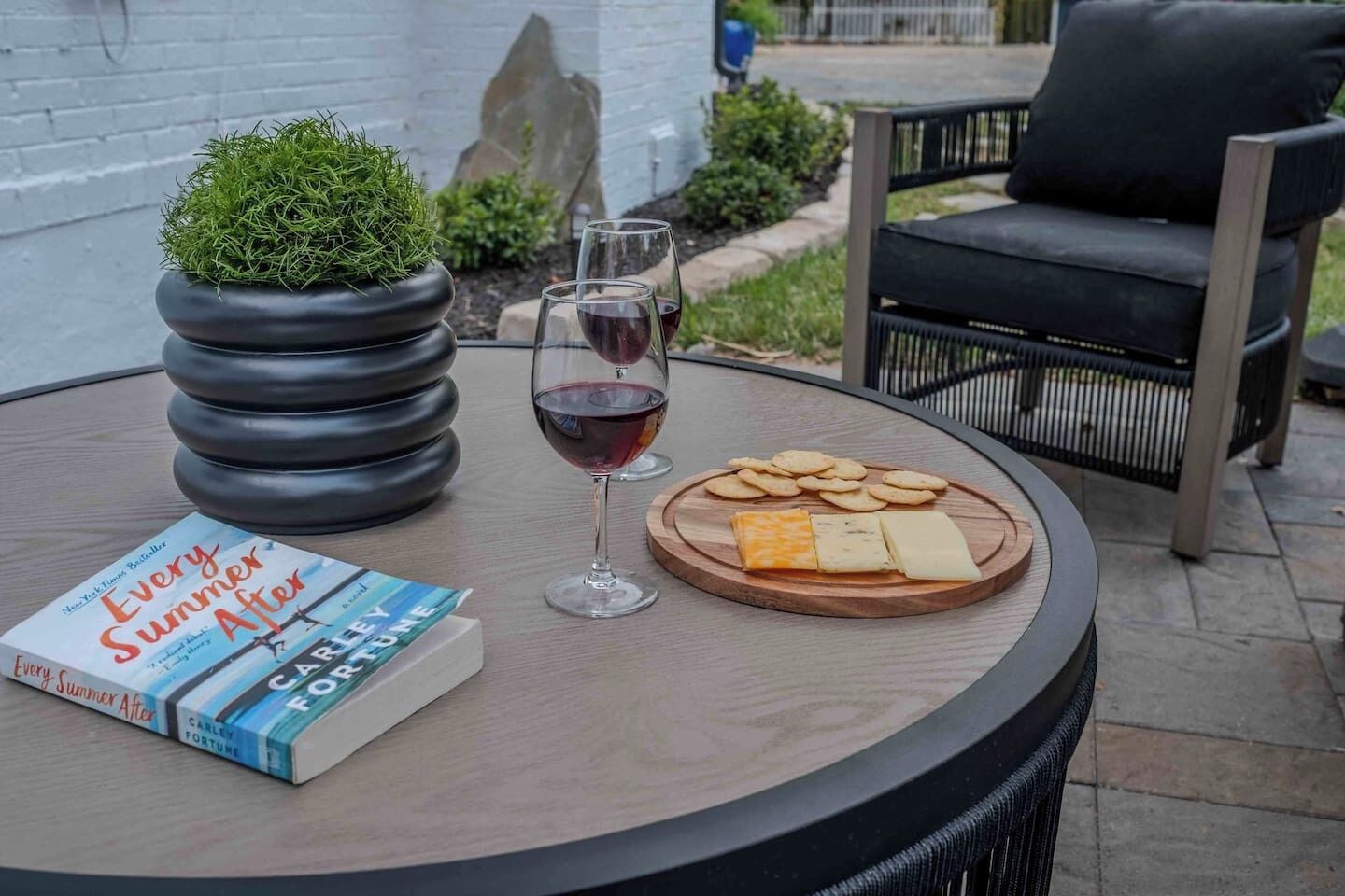 A round table with a book, two glasses of red wine, a potted plant, and a wooden board with cheese and crackers.