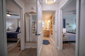 Un pasillo que conduce a un dormitorio con cama, cómoda y espejo. Se ve una puerta que da a otra habitación con escritorio y silla.