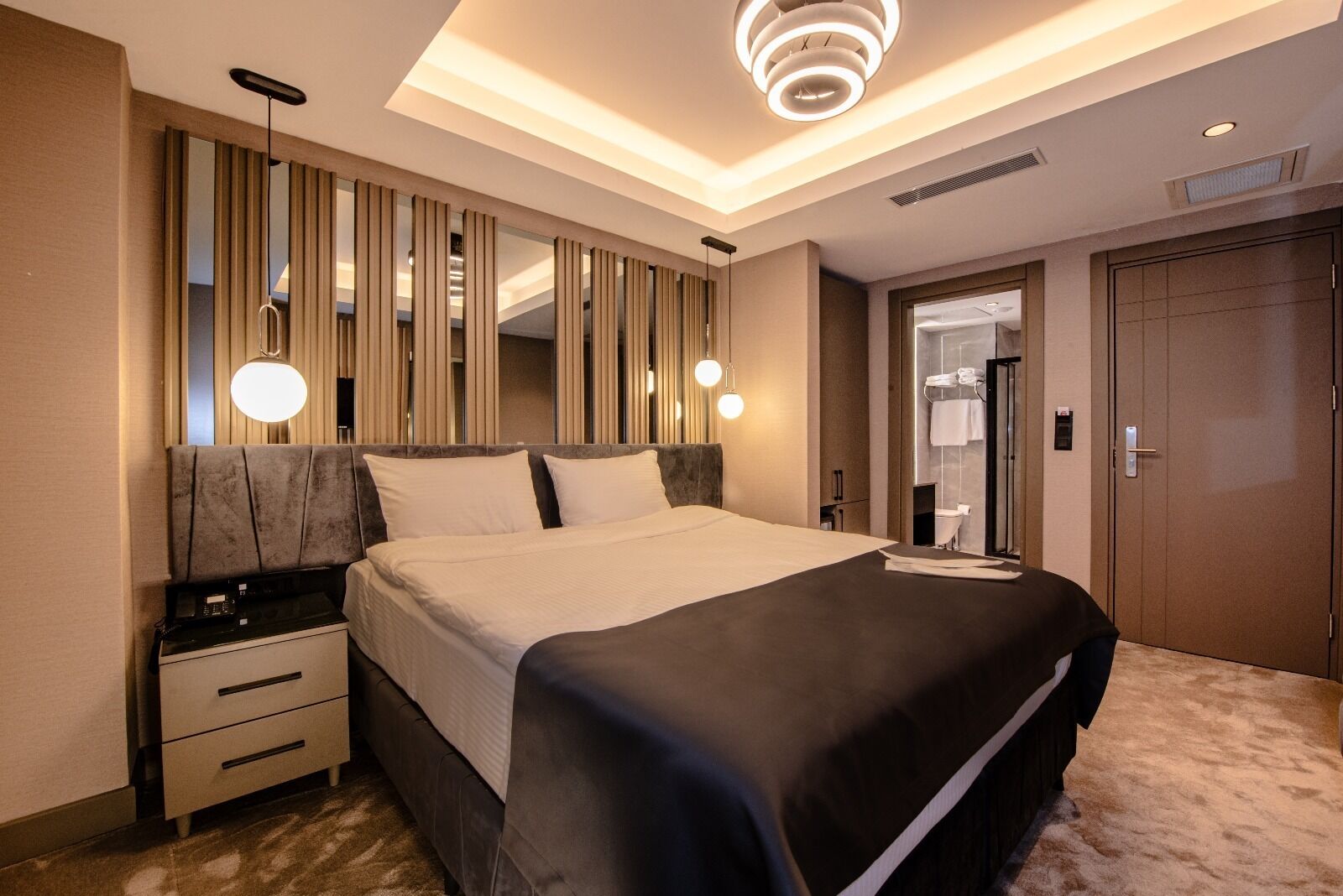 A modern hotel room with a large bed, bedside tables, a mirror, and a ceiling light fixture.