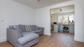 A modern living room with a grey sectional sofa, a washing machine, and a kitchen area with a window. - Spacious 3BDR Home with Workspace in Prime Spot (Greater London)