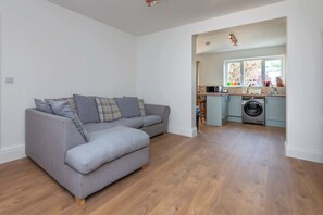 A modern living room with a grey sectional sofa, a washing machine, and a kitchen area with a window.