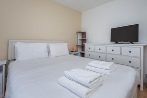A hotel room with a bed, a TV, a nightstand, a dresser, and a shelf. - Spacious 3BDR Home with Workspace in Prime Spot (Greater London)