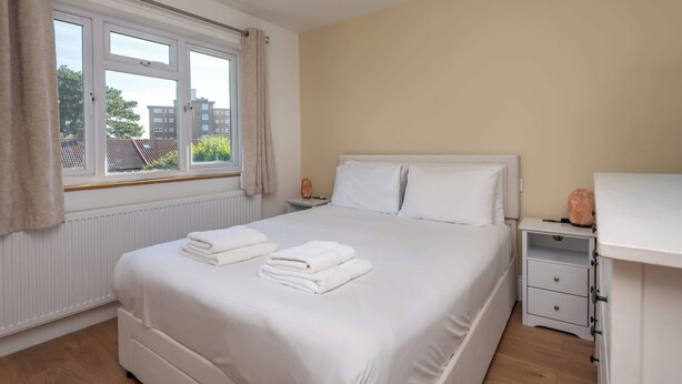 A neatly made bed with white linens, a bedside table with a lamp, and a window with curtains. - Spacious 3BDR Home with Workspace in Prime Spot (Greater London)