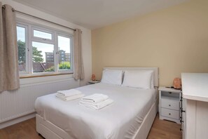 A neatly made bed with white linens, a bedside table with a lamp, and a window with curtains. - Spacious 3BDR Home with Workspace in Prime Spot (Greater London)