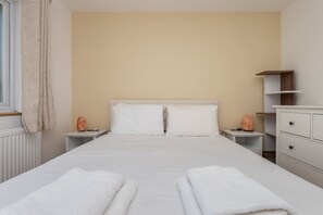 A neatly made bed with white linens, two bedside tables with decorative items, a white dresser with drawers, and a window with curtains.
