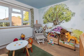 A child's playroom with a large window, a chair, a table, and a bookshelf filled with toys. - Spacious 3BDR Home with Workspace in Prime Spot (Greater London)