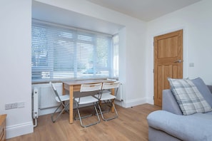 A room with a wooden table, three white chairs, a grey sofa, a wooden door, and a window with blinds. - Spacious 3BDR Home with Workspace in Prime Spot (Greater London)