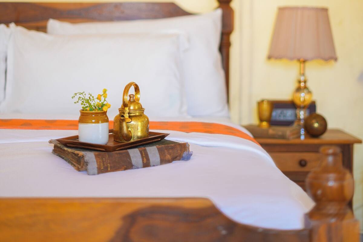 Una cama bien tendida con ropa de cama blanca, un cabecero de madera y una mesita de noche con una lámpara y un reloj.