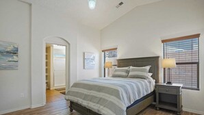 Een slaapkamer met een bed, nachtkastjes, een raam met jaloezieën, een deur en een gang.