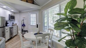 Una cocina compacta con armarios blancos, un microondas y un lavamanos. Hay una mesa de comedor con sillas blancas y una planta en maceta.