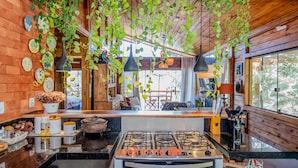 A rustic kitchen with a gas stove, wooden walls, hanging plants, and a dining area with a table and chairs.