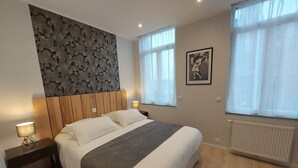 A bedroom with a bed, nightstands, a wall-mounted light, a radiator, and a window with sheer curtains. - B As in... Forest walk (Saint-Amand-les-Eaux)