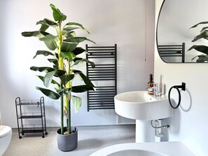 Un baño moderno con un lavabo blanco, un perchero negro, una planta en maceta y una estantería metálica.