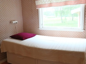 A single bed with a white mattress and a purple pillow, a window with a patterned curtain, and a wall with a floral wallpaper pattern.