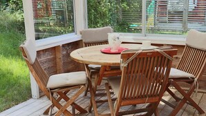 Una terraza de madera con una mesa redonda y cuatro sillas, rodeada por una valla de madera y una ventana con persianas.